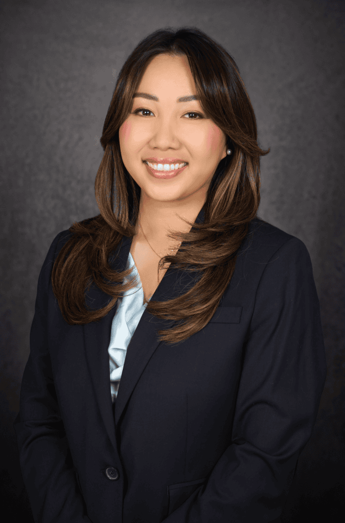 A woman with long brown hair, wearing a navy blazer and light blue blouse, smiles at the camera against a dark, textured background.