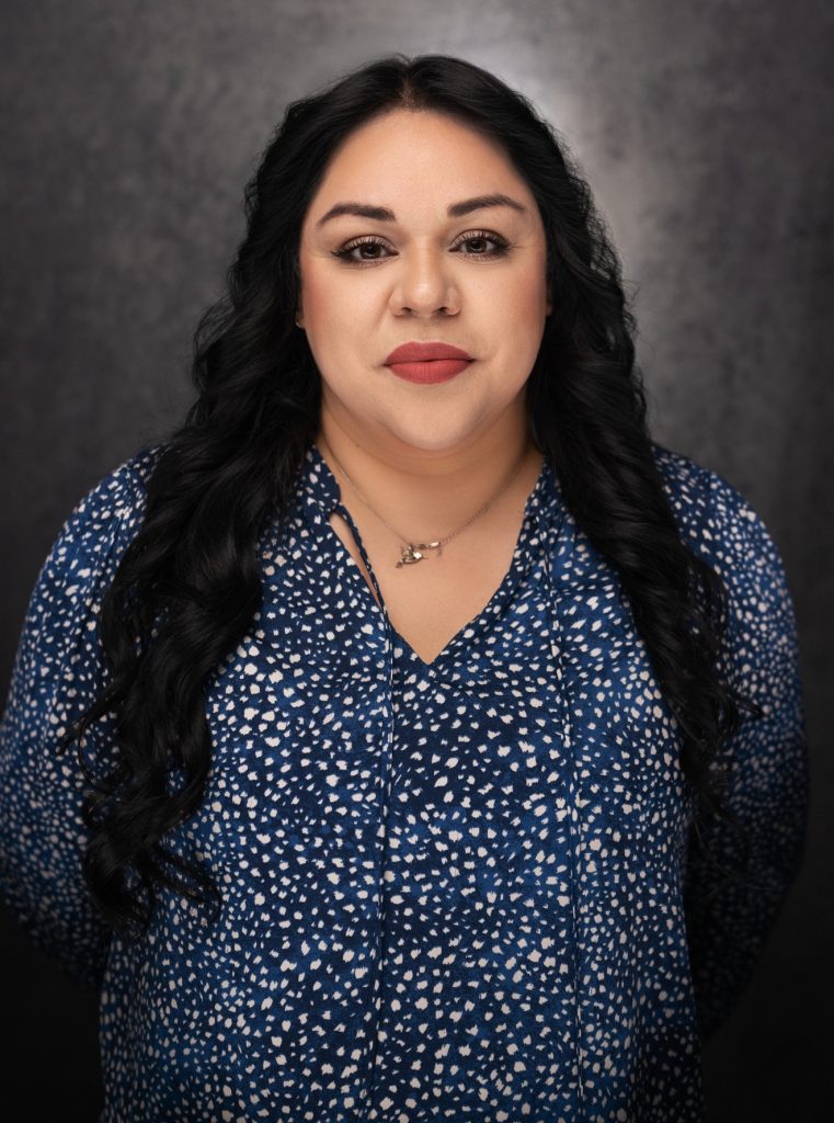 Woman with long dark wavy hair, wearing a blue patterned blouse and a necklace, stands against a dark textured background, looking directly at the camera with a neutral expression.