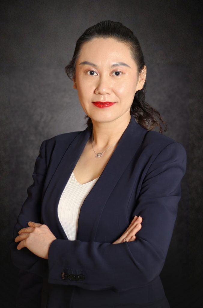 A woman in a navy blazer and white top stands with arms crossed, smiling confidently against a dark, textured background.