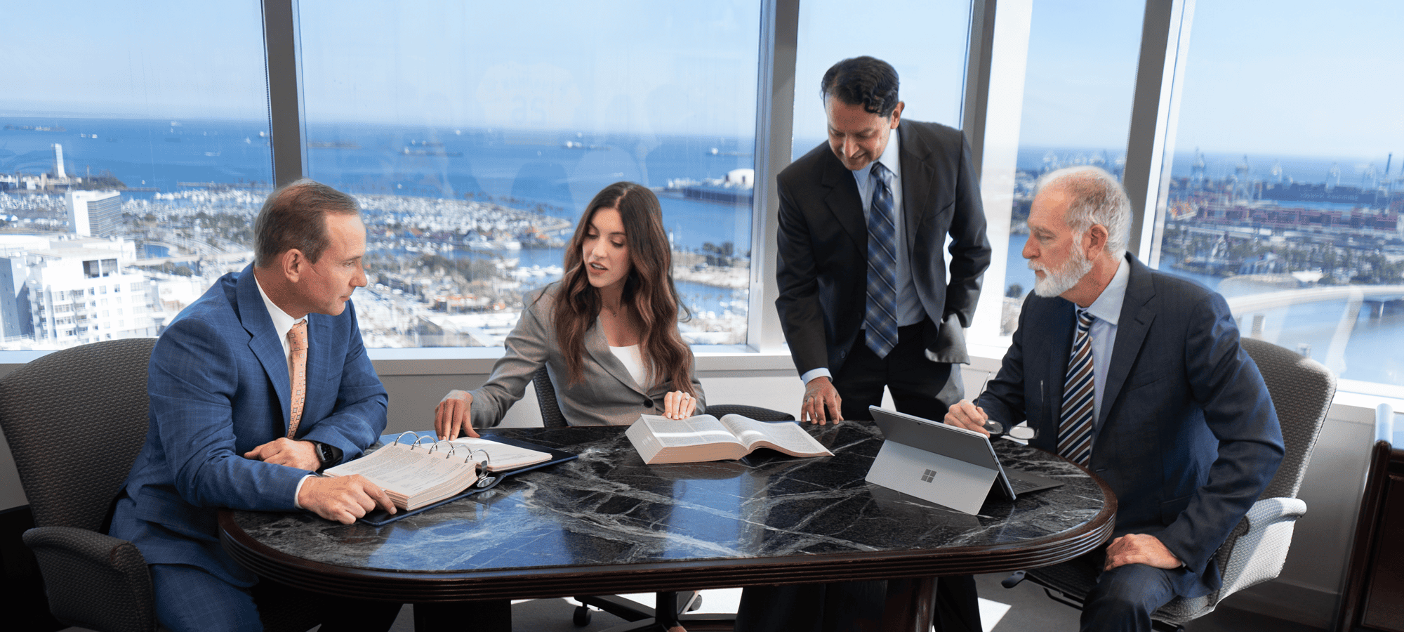 Four business people in suits sit and stand around a conference table with open books and a laptop, discussing work in a high-rise office with large windows overlooking a harbor and cityscape.